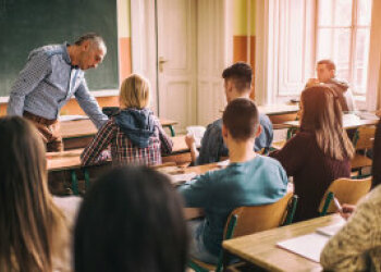 Blick von hinten in ein Klassenzimmer mit sitzenden Schülerinnen und Schülern. Vorne steht ein Mann.